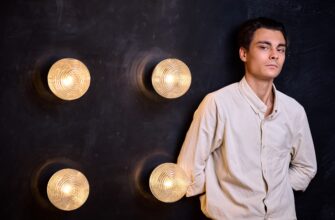 На изображении человек в светлой рубашке стоит у тёмной стены с четырьмя круглыми светильниками. Светильники излучают тёплый свет.