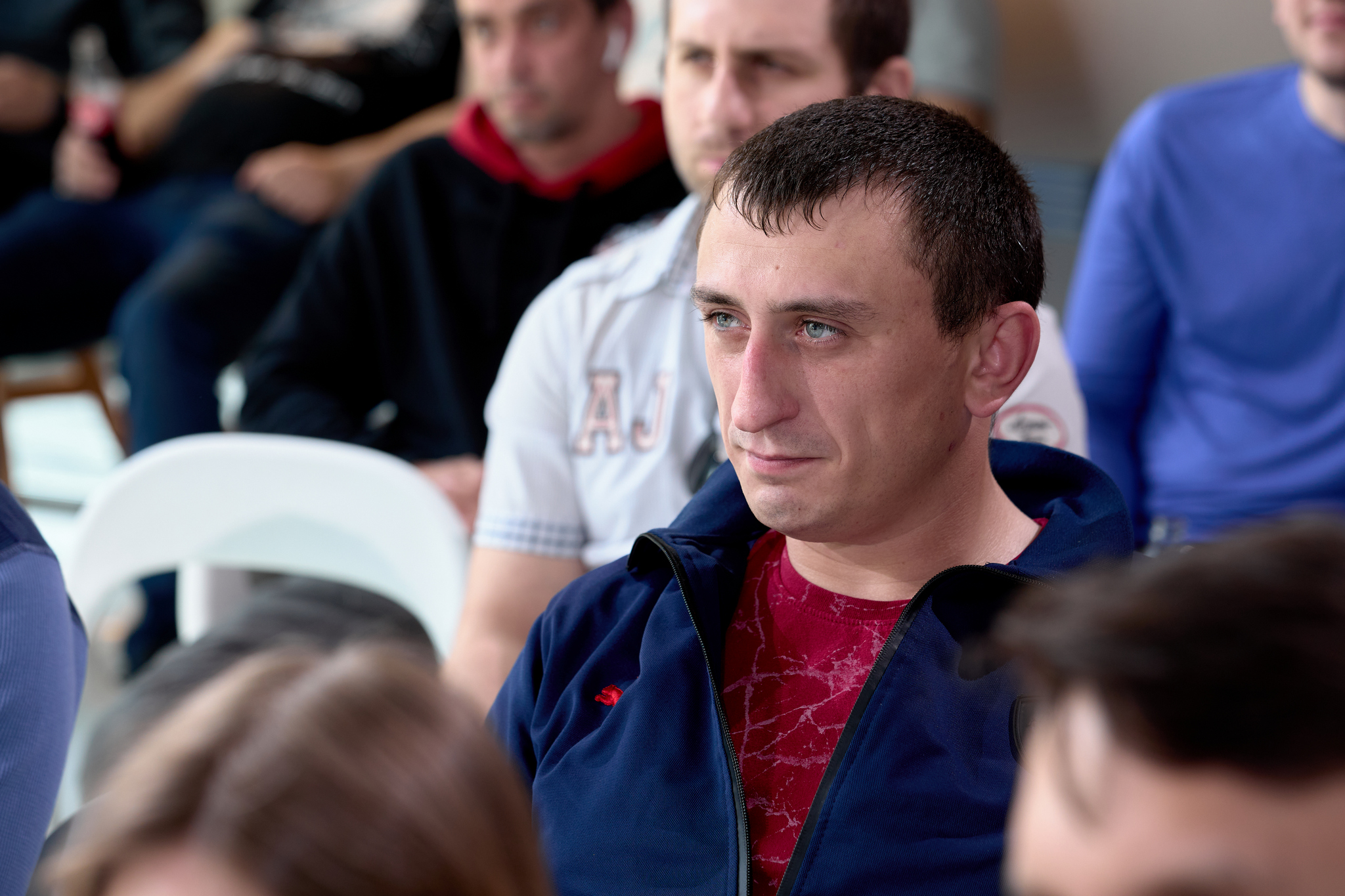На изображении группа людей сидит в помещении. На переднем плане мужчина в синей куртке и красной футболке.