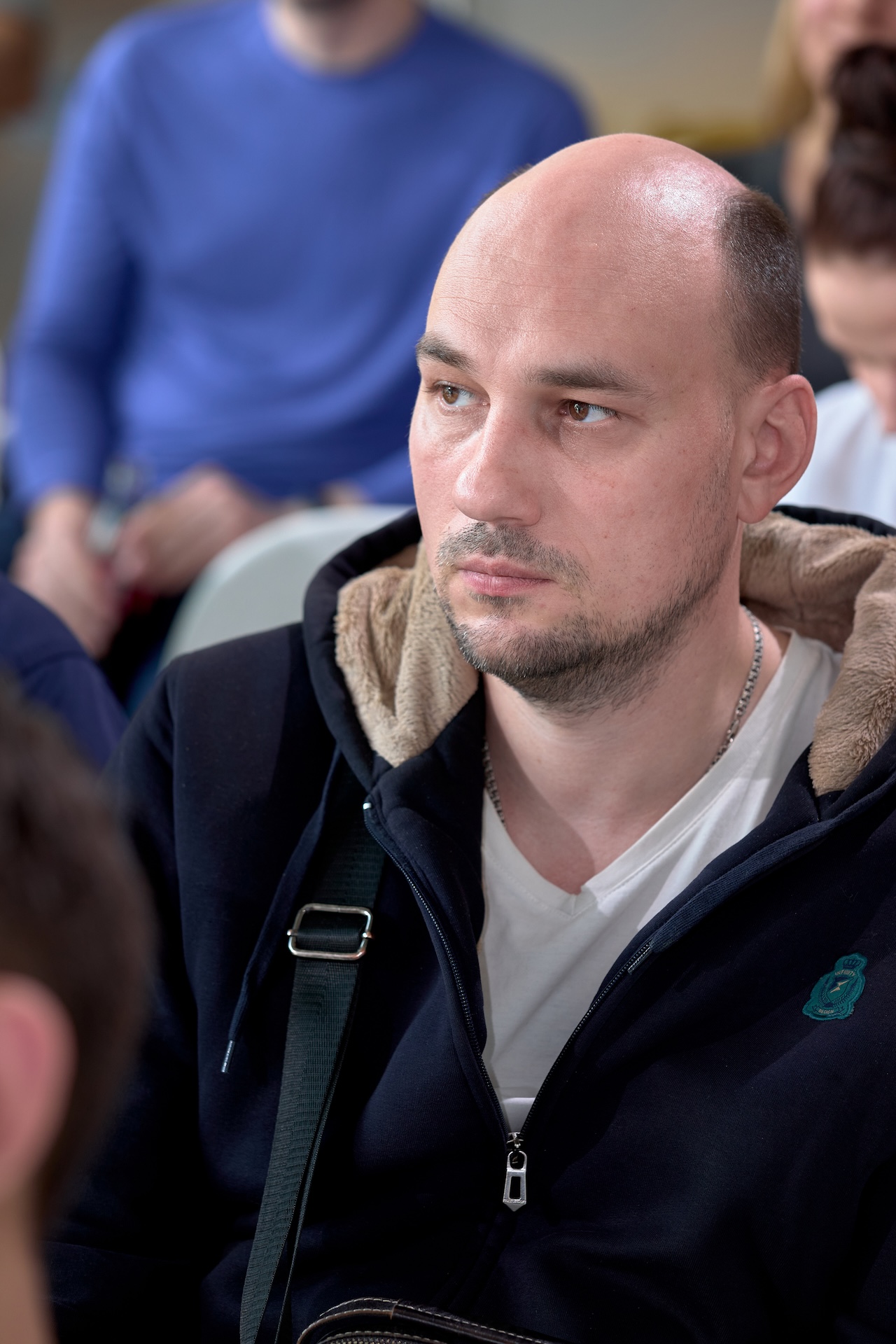 На изображении мужчина с бритой головой и бородой. Он одет в тёмную куртку с капюшоном на меховой подкладке и белую футболку.