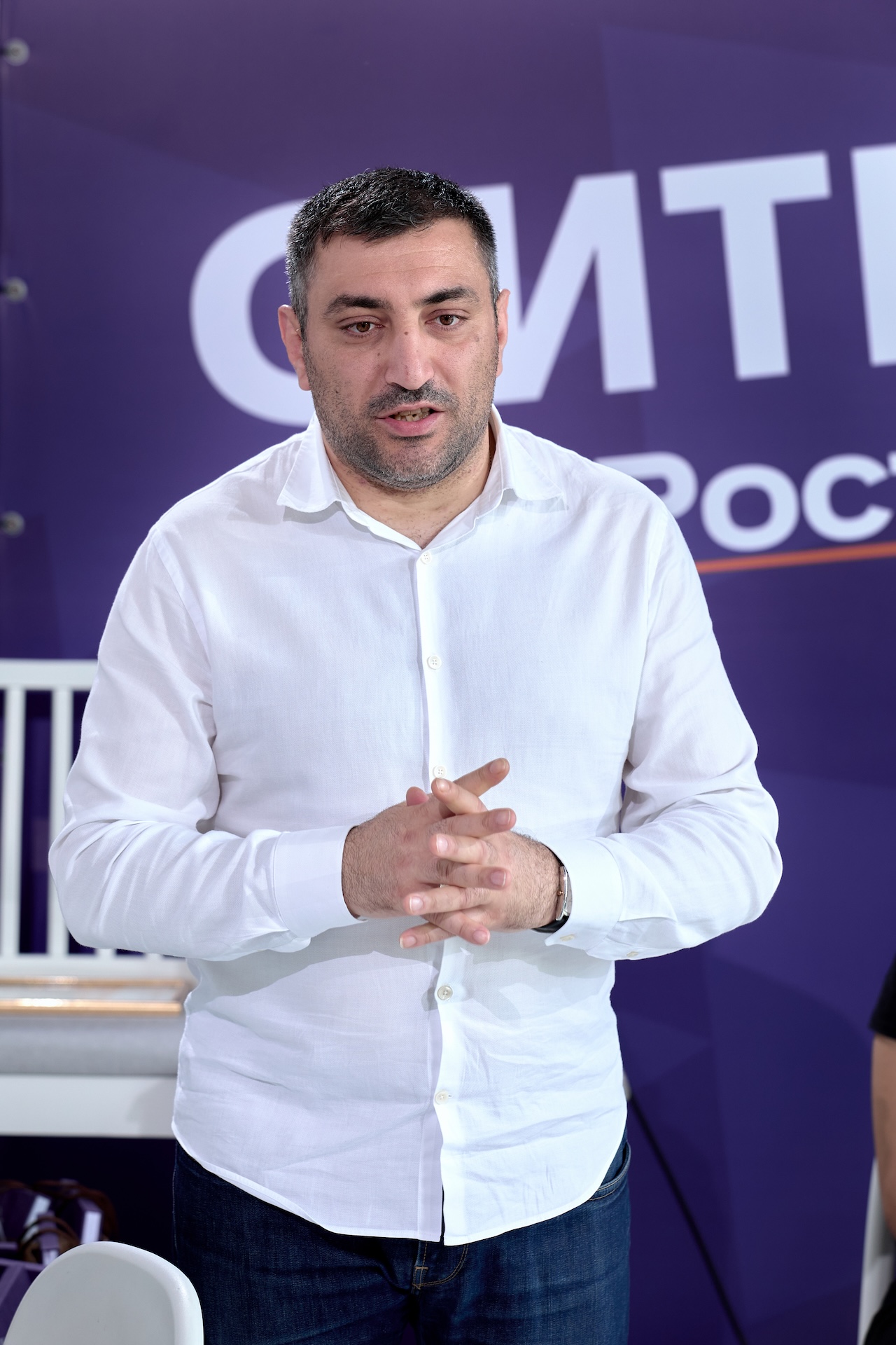 Мужчина в белой рубашке и джинсах стоит на фоне фиолетового баннера с белыми буквами. Он жестикулирует руками.