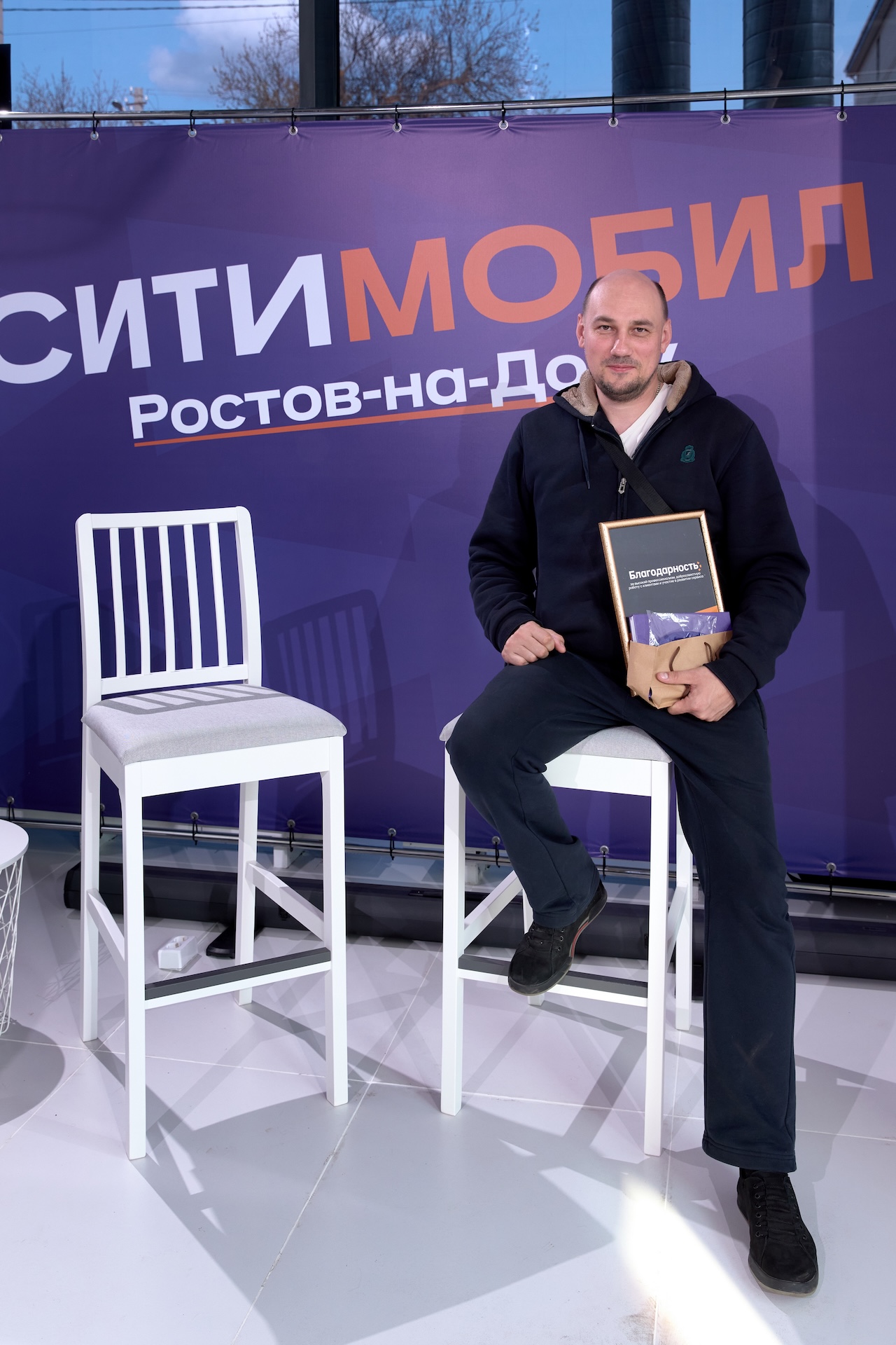 Мужчина в тёмной одежде сидит на белом стуле, держа в руках коробку. На фоне фиолетовый баннер с надписью «СитиМобил Ростов-на-Дону».