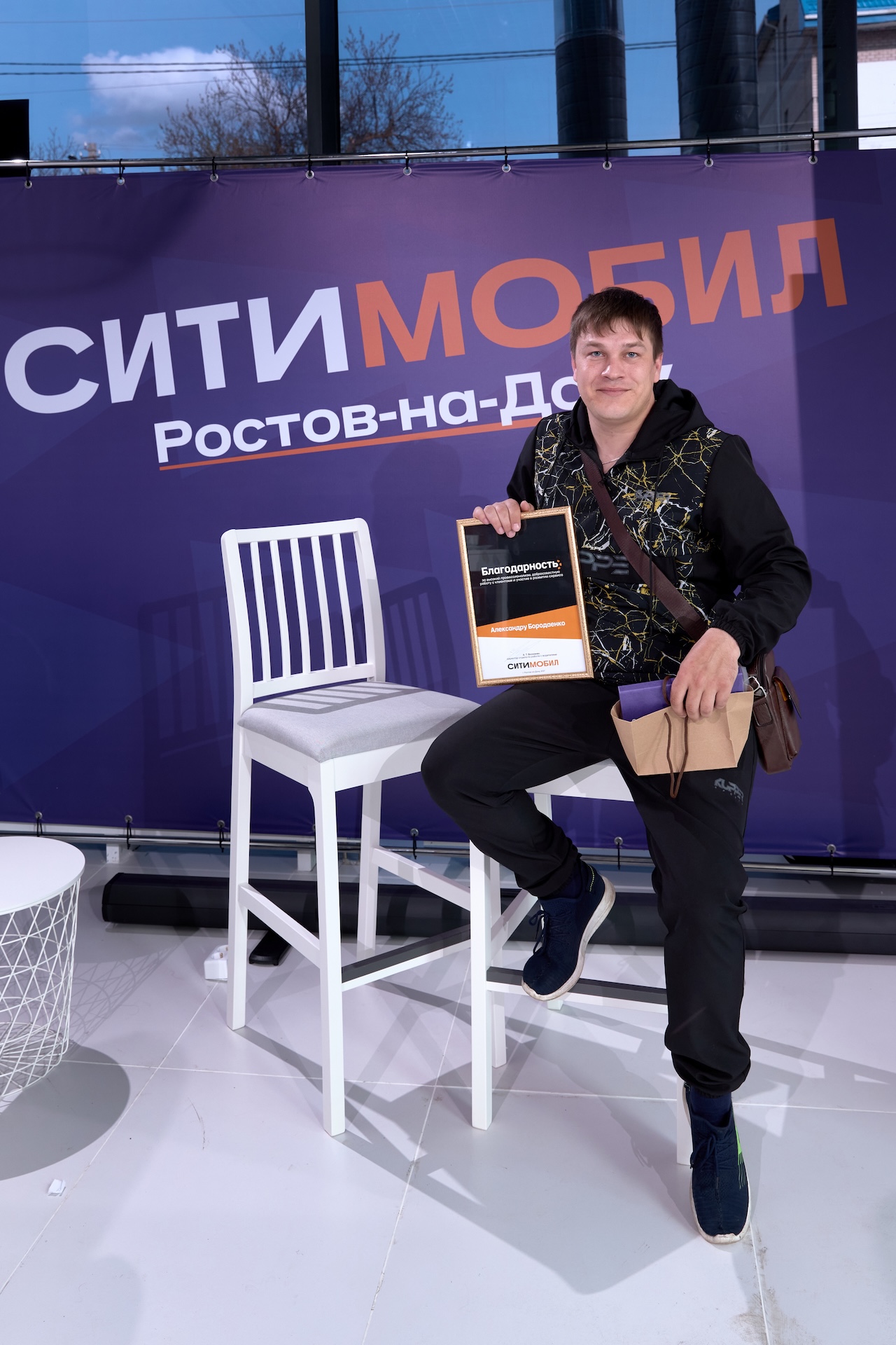 Человек сидит на стуле, держит коробку и сумку. На фоне фиолетовая панель с надписью «Ситимобил Ростов-на-Дону».