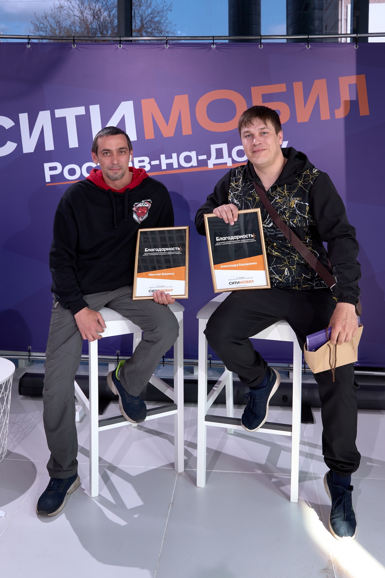 На изображении двое мужчин сидят на белых табуретах, держат сертификаты и улыбаются. На фоне баннер с надписью «СИТИМОБИЛ Ростов-на-Дону».