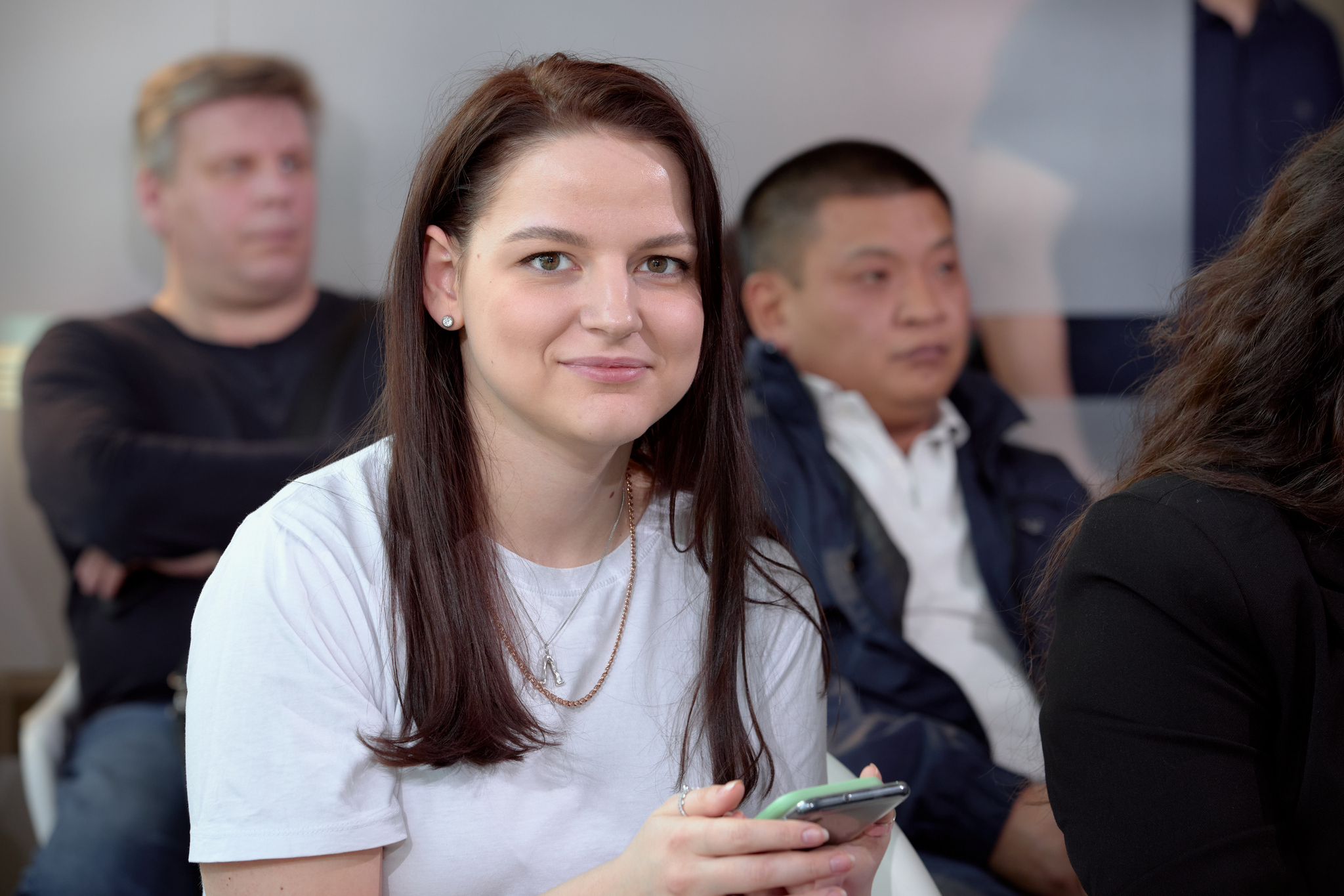 На изображении молодая женщина в белой футболке с длинными каштановыми волосами. Она держит телефон и улыбается. Вокруг неё сидят люди.