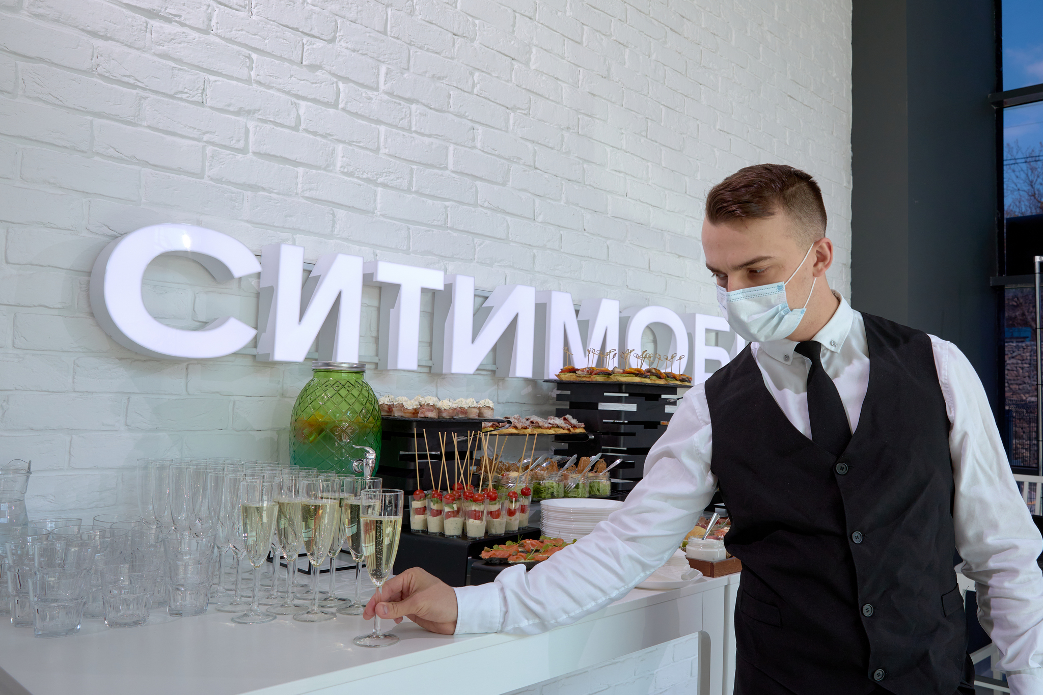 На изображении мужчина в маске и костюме стоит у стойки с напитками. На стене надпись «Ситимобил». На стойке бокалы и закуски.