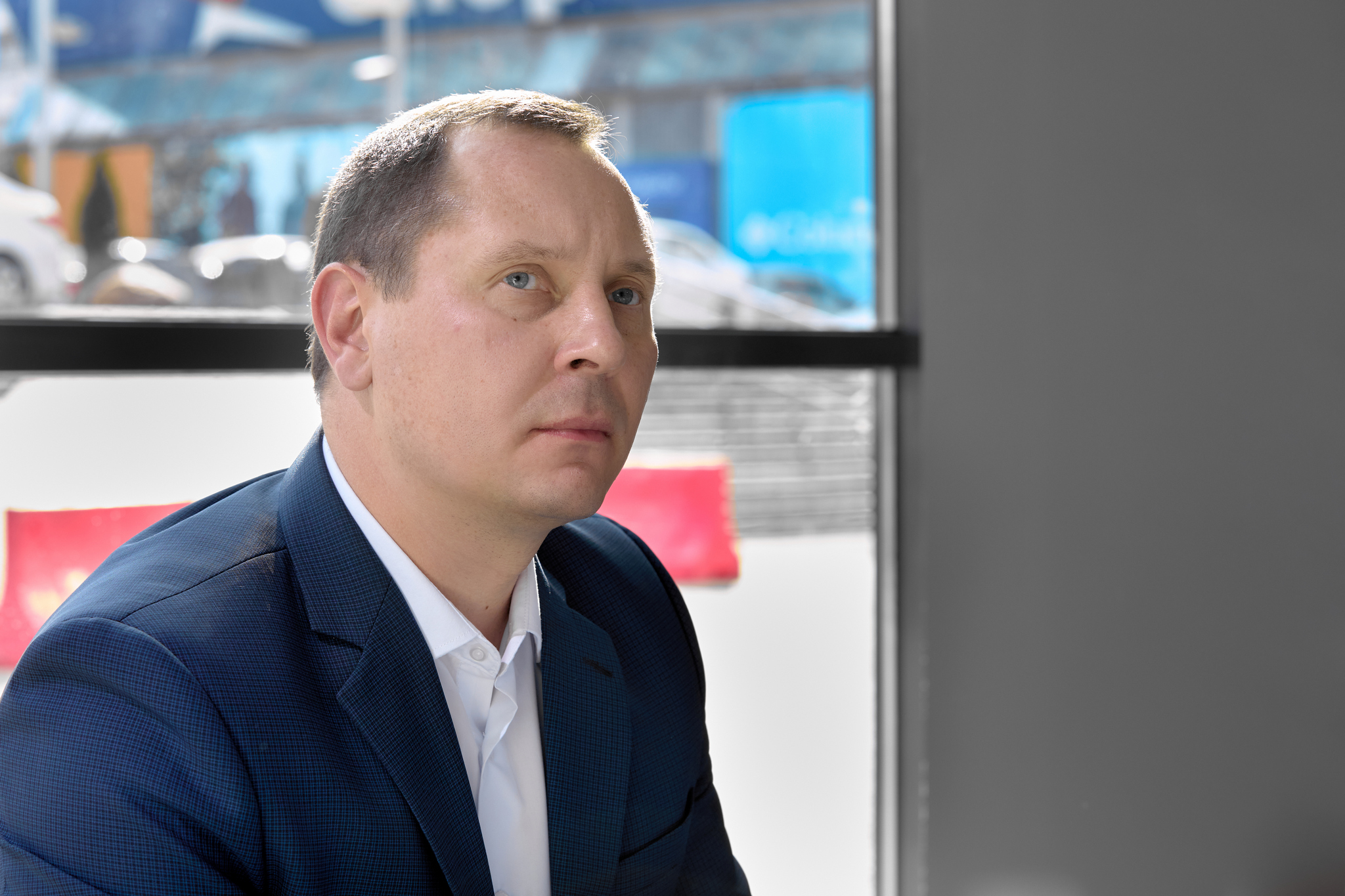 На изображении мужчина в костюме, белой рубашке. Он сидит в помещении с большими окнами, через которые видны машины и здания.