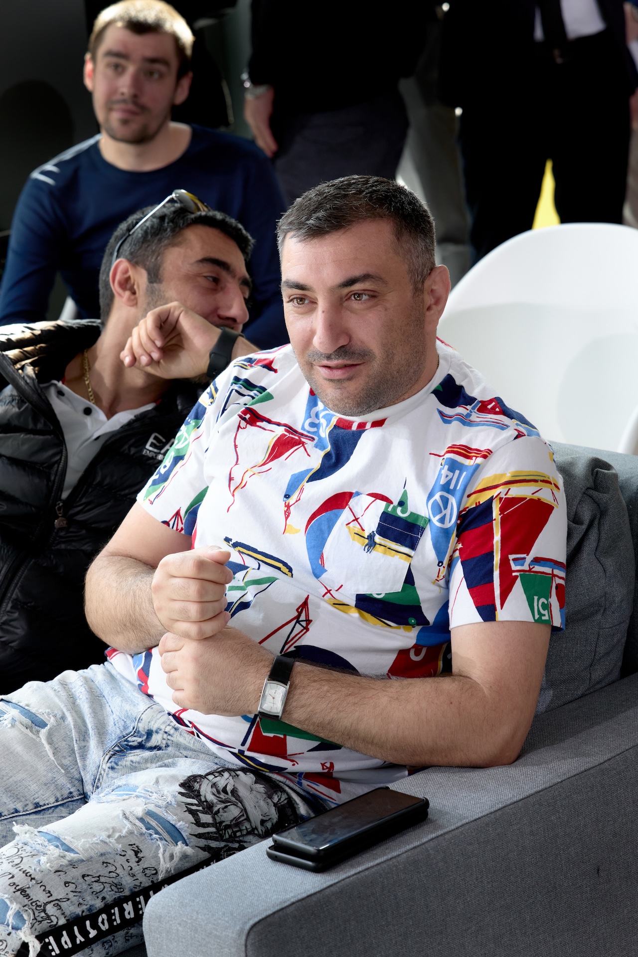 На изображении трое мужчин. Один сидит на диване в яркой футболке и светлых брюках, рядом лежит телефон.