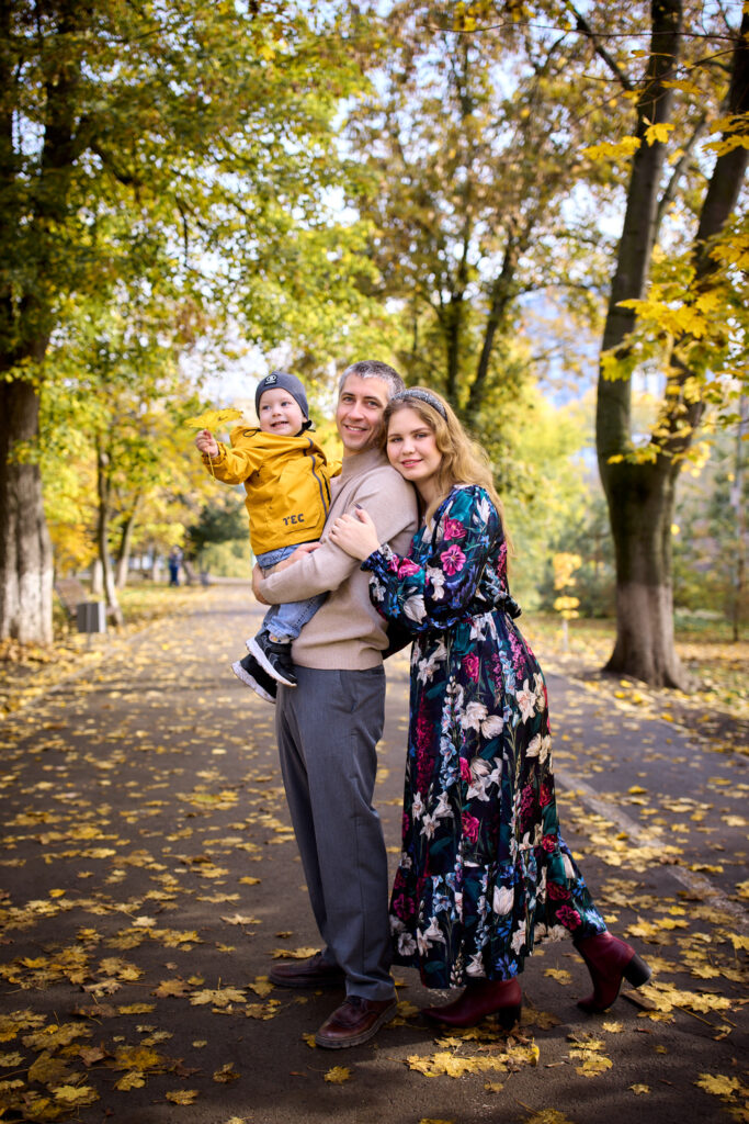 Семейная фотосессия на природе - мама, папа и ребёнок. Они стоят на дорожке в парке, окружённом деревьями с жёлтыми листьями.