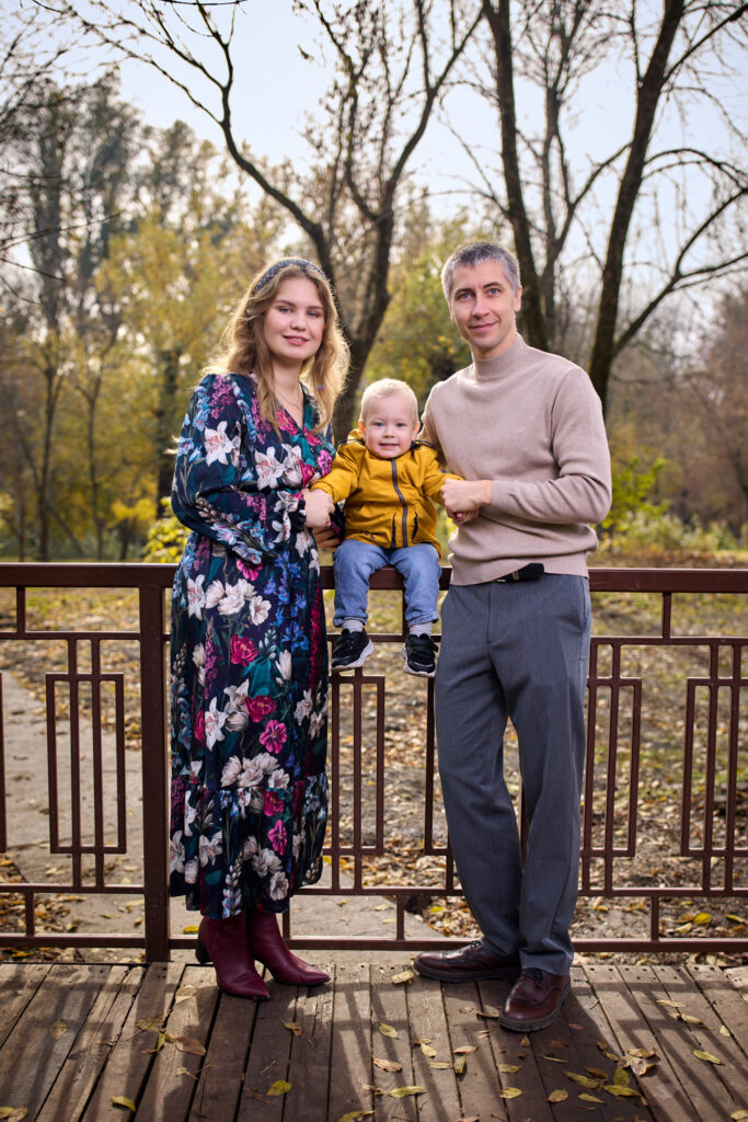 Семейная фотосессия на природе - мама, папа и ребёнок. Они стоят на дорожке в парке, окружённом деревьями с жёлтыми листьями.
