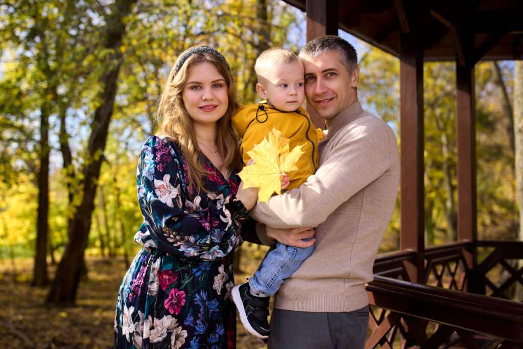 Семейная фотосессия на природе - женщина в платье с цветами, мужчина в свитере и ребёнок в жёлтой одежде. Они стоят на улице, на фоне деревьев и беседки.