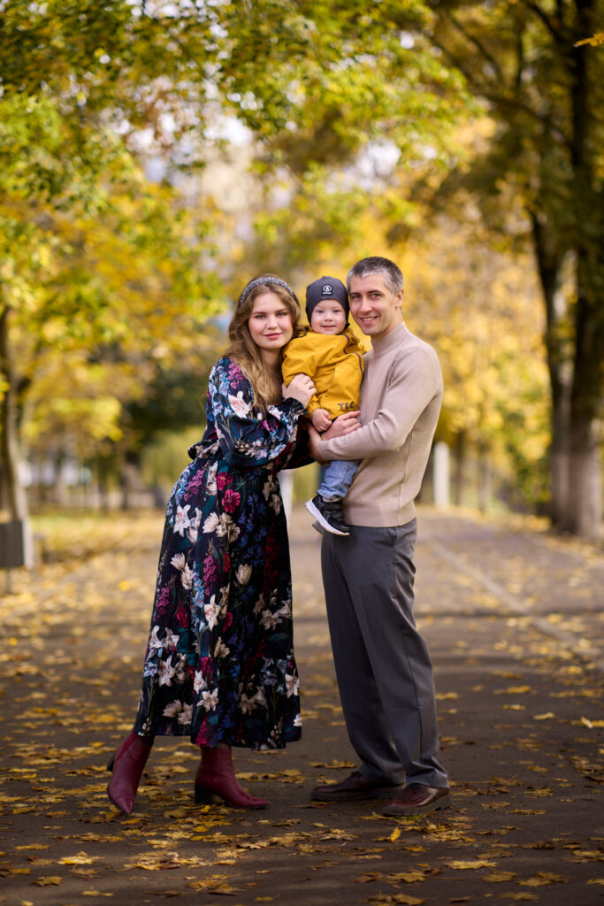 Семейная фотосессия на природе - мама, папа и ребёнок. Они стоят на дорожке в парке, окружённом деревьями с жёлтыми листьями.