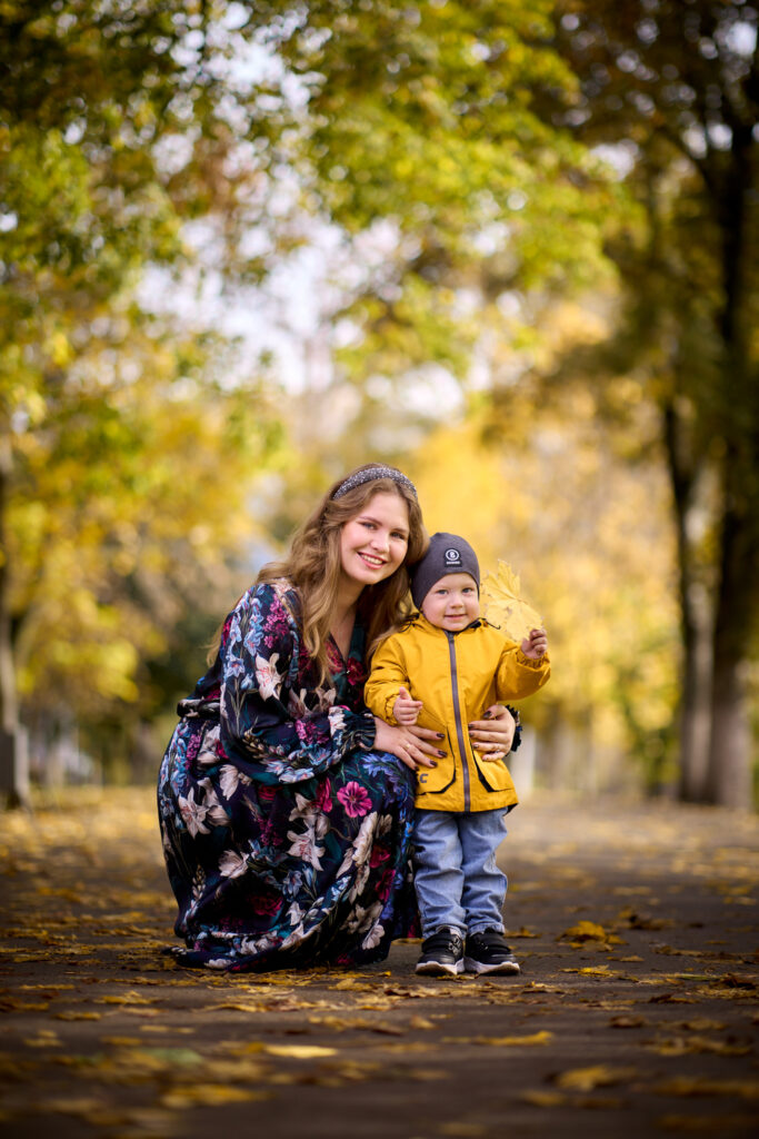 Семейная фотосессия на природе - мама в платье с цветами и сын в жёлтой куртке и шапке. Они стоят на дорожке в парке, вокруг деревья с жёлтыми листьями.