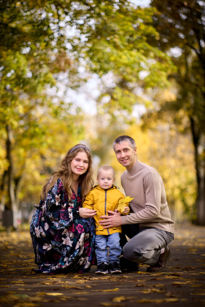 Семейная фотосессия на природе -семья из трёх человек: отец, мать и сын. Они сидят на земле в парке, окружённом деревьями с жёлтыми листьями.