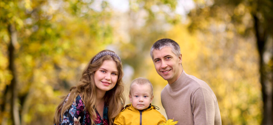 Семейная фотосессия на природе -семья из трёх человек: отец, мать и сын. Они сидят на земле в парке, окружённом деревьями с жёлтыми листьями.