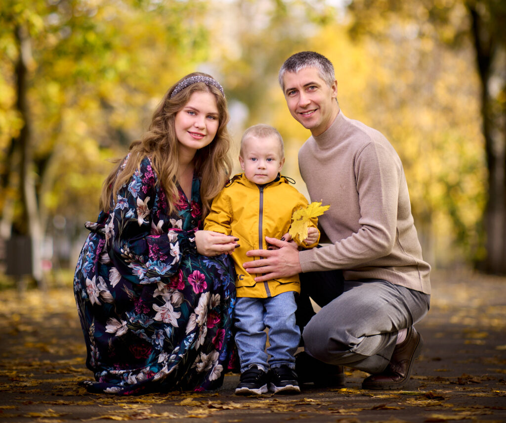 Семейная фотосессия на природе -семья из трёх человек: отец, мать и сын. Они сидят на земле в парке, окружённом деревьями с жёлтыми листьями.