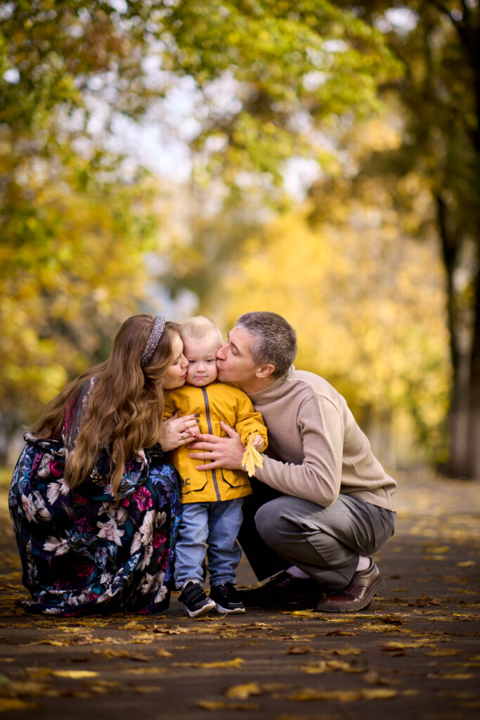 Семейная фотосессия на природе -семья из трёх человек: отец, мать и сын. Они сидят на земле в парке, окружённом деревьями с жёлтыми листьями.
