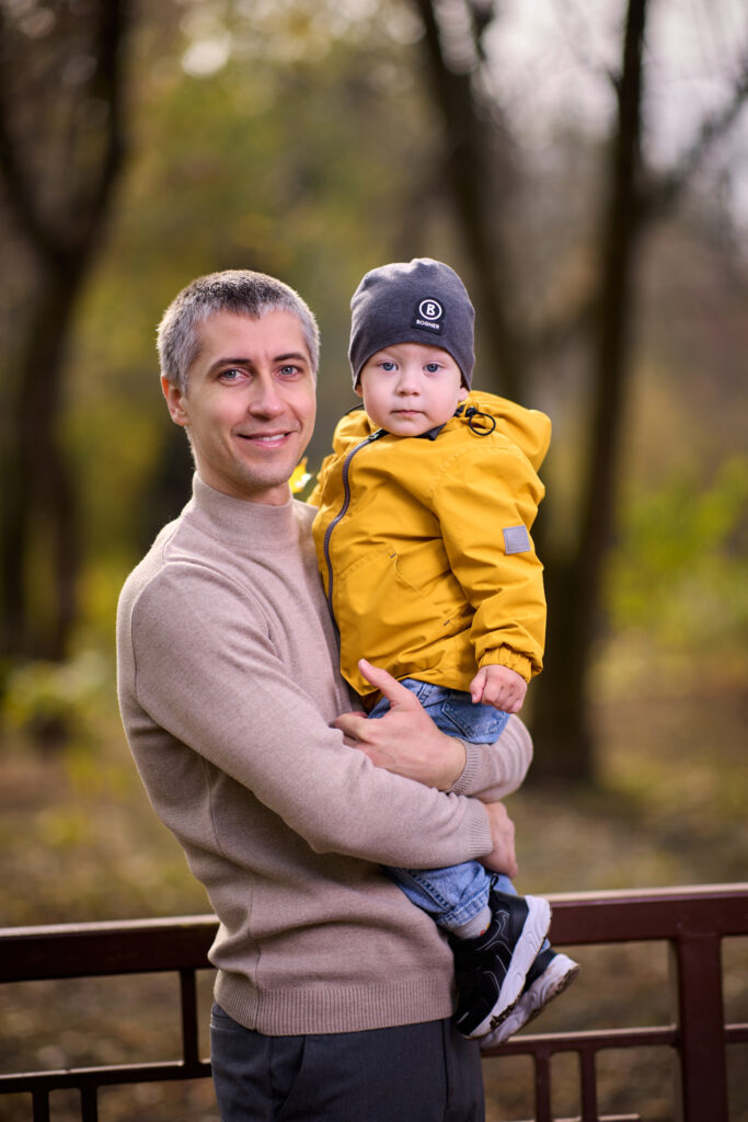 Семейная фотосессия на природе - отец держит сына на руках. Ребёнок в жёлтой куртке и серой шапке. Фон размытый, с деревьями.