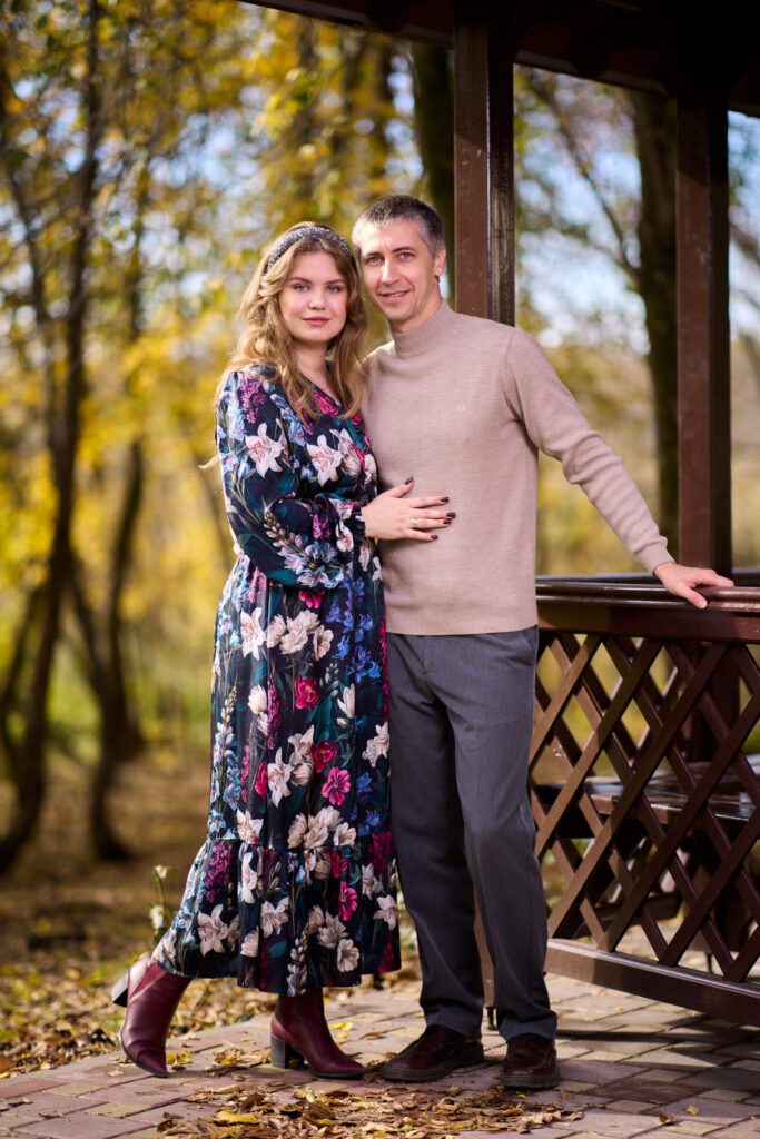 Семейная фотосессия на природе - мужчина и женщина стоят в парке, обнявшись. Вокруг деревья и опавшие листья.